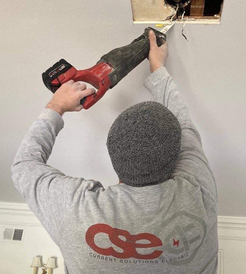 Electrician from Current Solutions Electric using a reciprocating saw to perform electrical work on a ceiling, demonstrating professional service and expertise in Kansas City.