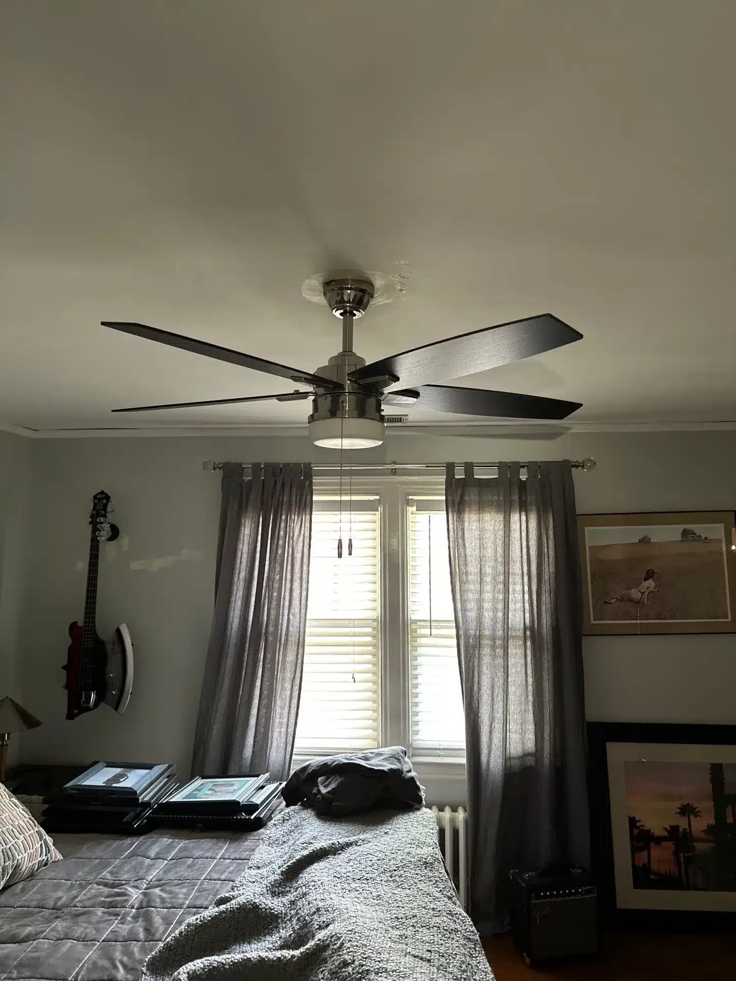 Ceiling fan with modern design and dark blades installed in a bedroom, showcasing a cozy interior with gray curtains and framed artwork, relevant to ceiling fan installation services in Kansas City, MO.