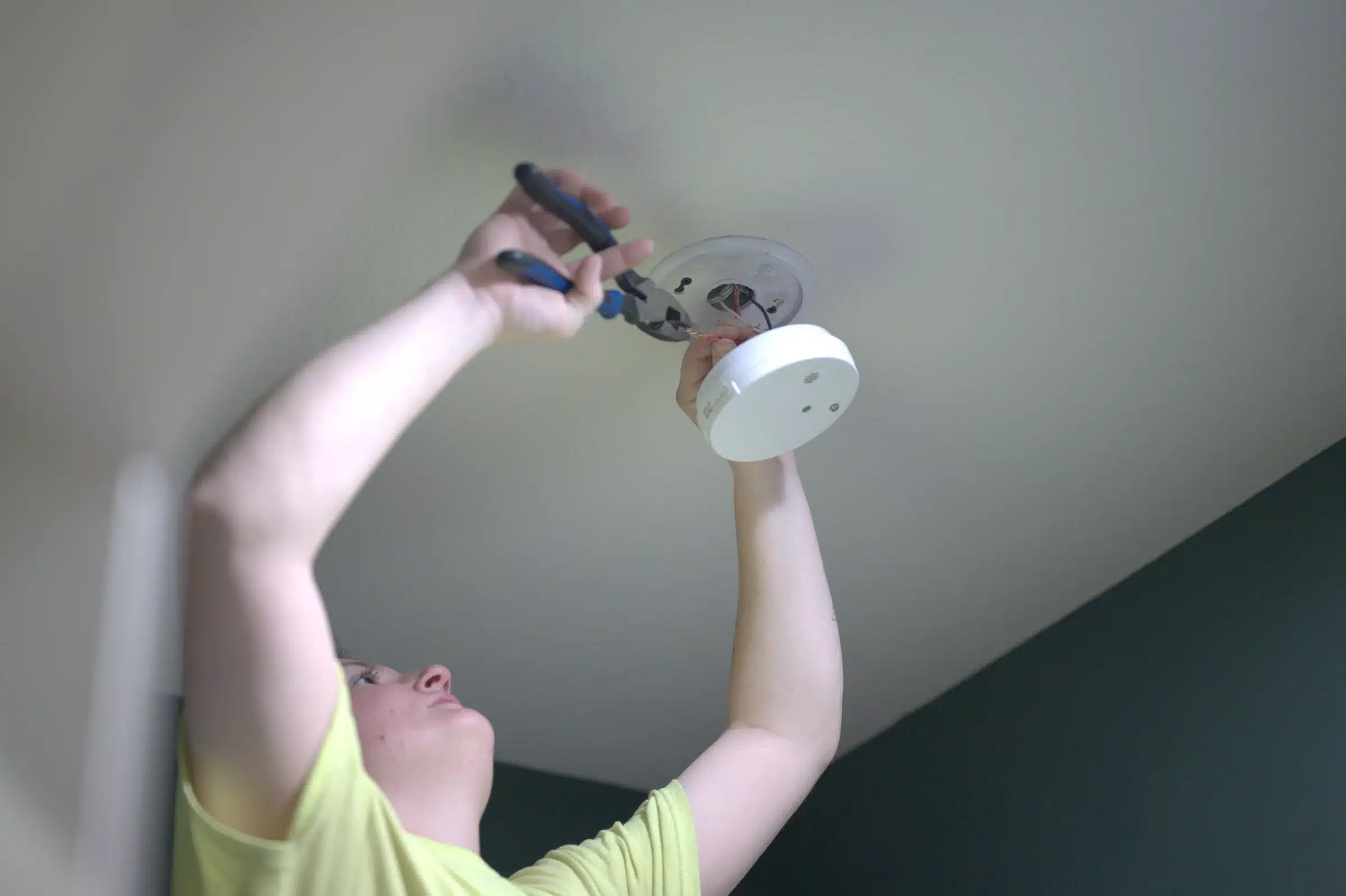 Person installing a smoke detector on a ceiling, using pliers, emphasizing professional smoke detector installation services in Kansas City, MO.