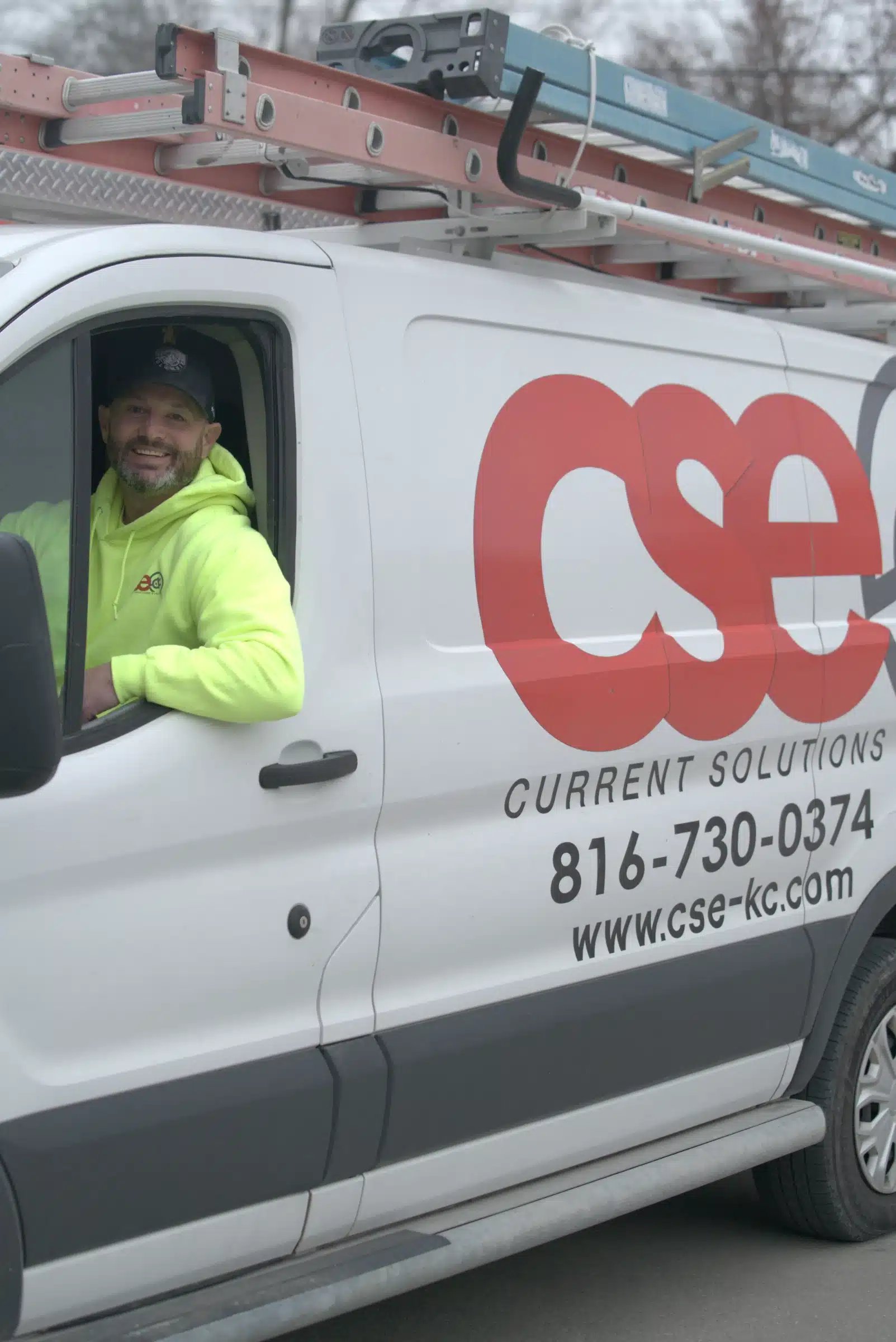 Man in a bright yellow hoodie smiling from the driver's seat of a Current Solutions Electric van, showcasing the company's branding and services related to electric vehicle charger installation in Kansas City, MO.