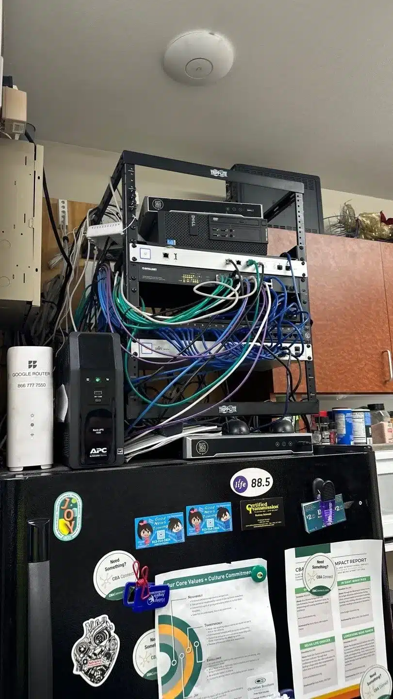Rack of networking equipment with cables, Google router, and UPS on top of a refrigerator, illustrating home automation technology setup in a Kansas City residence.