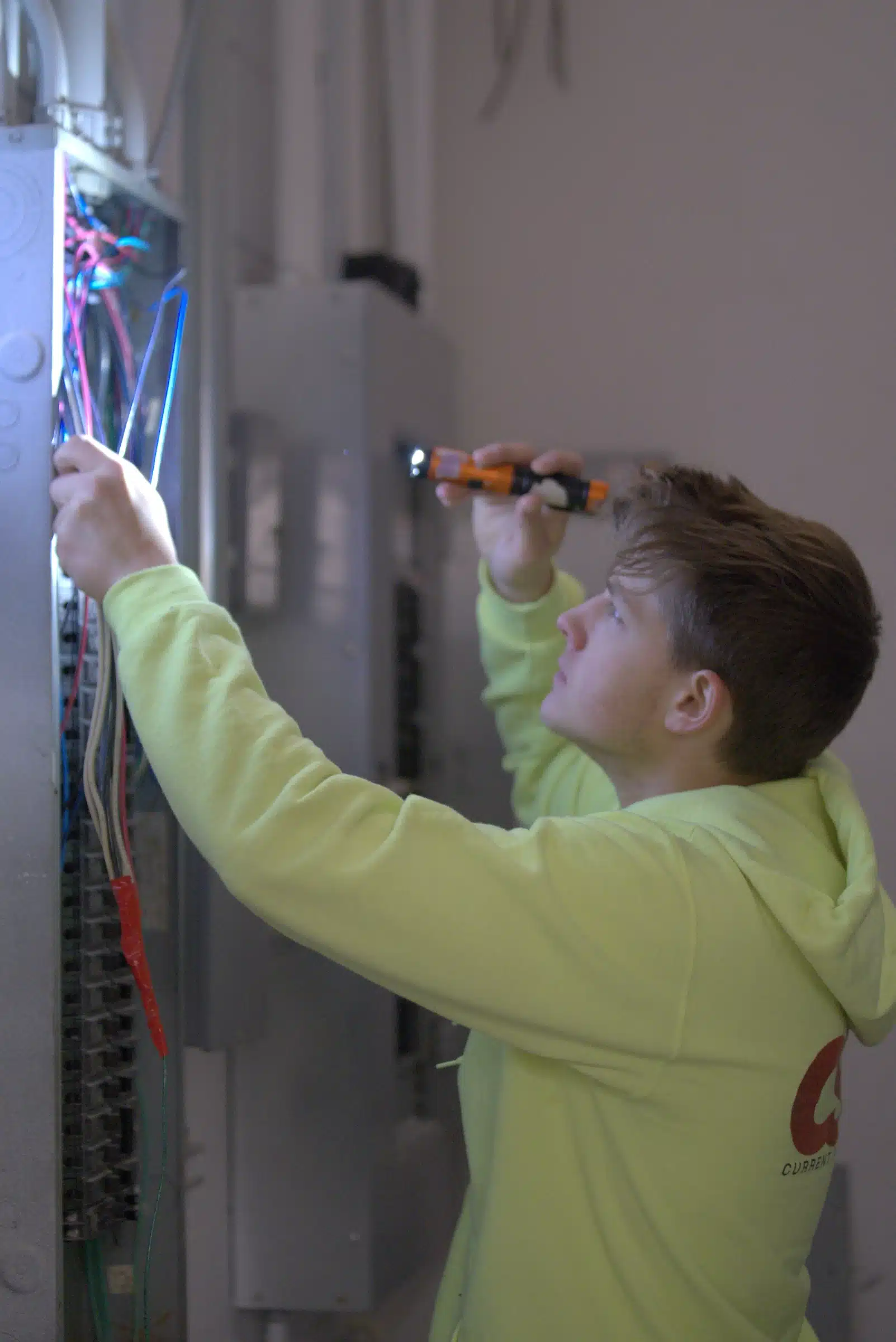 Electrician in bright yellow hoodie working on electrical panel, inspecting wires and connections for safe outlet and switch installation in Kansas City, MO.